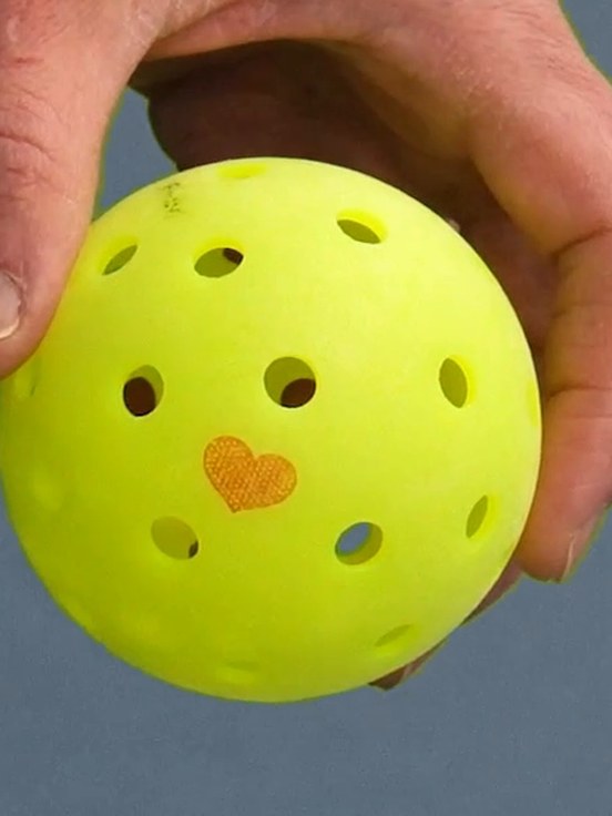 A hand holding a pickleball stamped with a Dink Ink heart, on a pickleball court